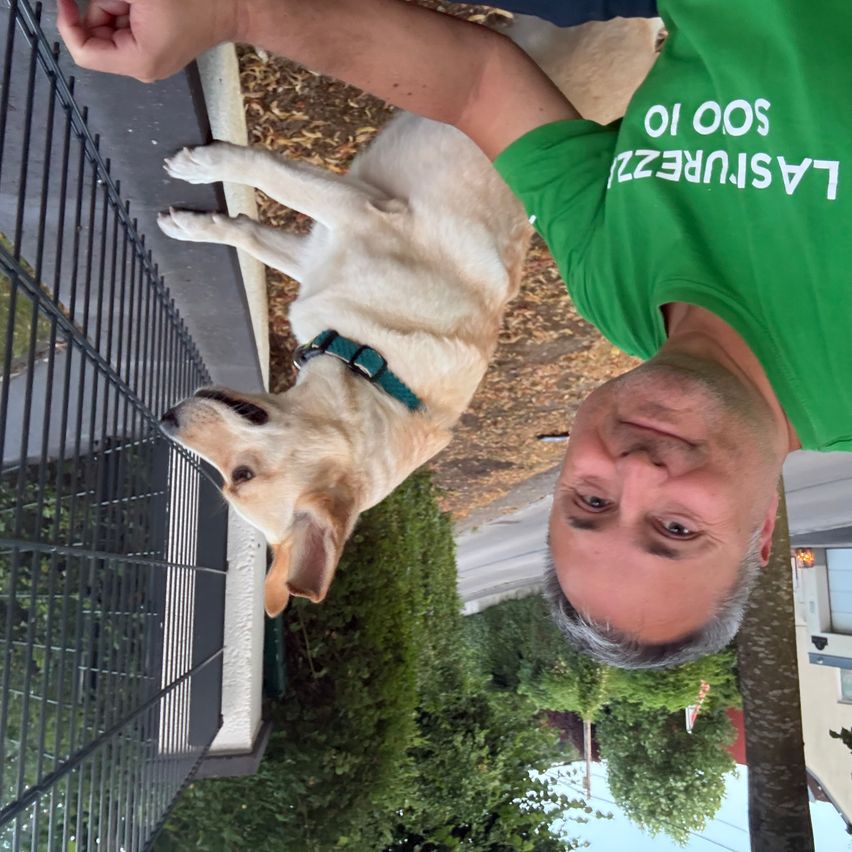 Ein Mann in einem grünen T-Shirt mit dem Wort 'LASER' darauf hält einen Hund, der auf seinem Arm steht. Der Hund schaut nach oben.
