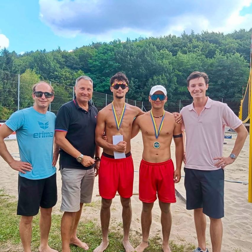 Beachvolleyball-Sieger mit Medaillen und Funktionären auf Sandplatz mit Wald im Hintergrund.