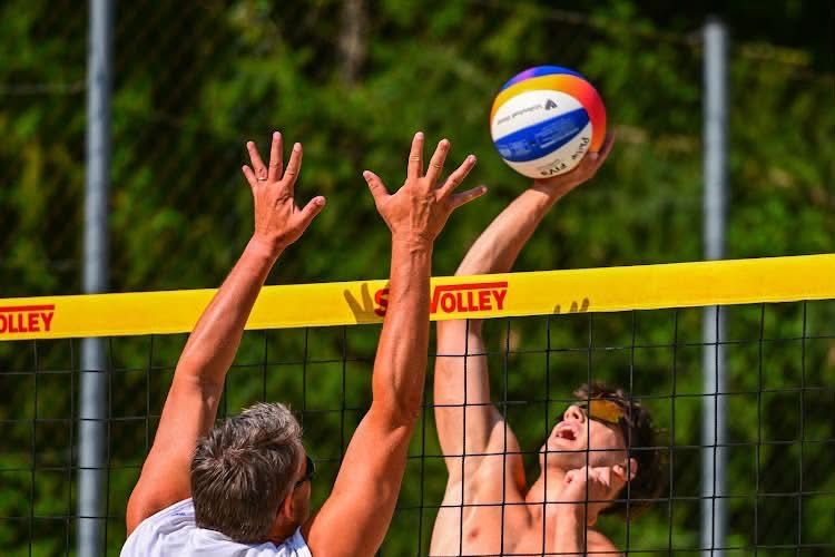 Zwei Männer spielen Volleyball, einer springt, um den Ball zu schlagen, der andere blockt mit erhobenen Händen, vor einem Netz mit dem Wort 'Volley' darauf.