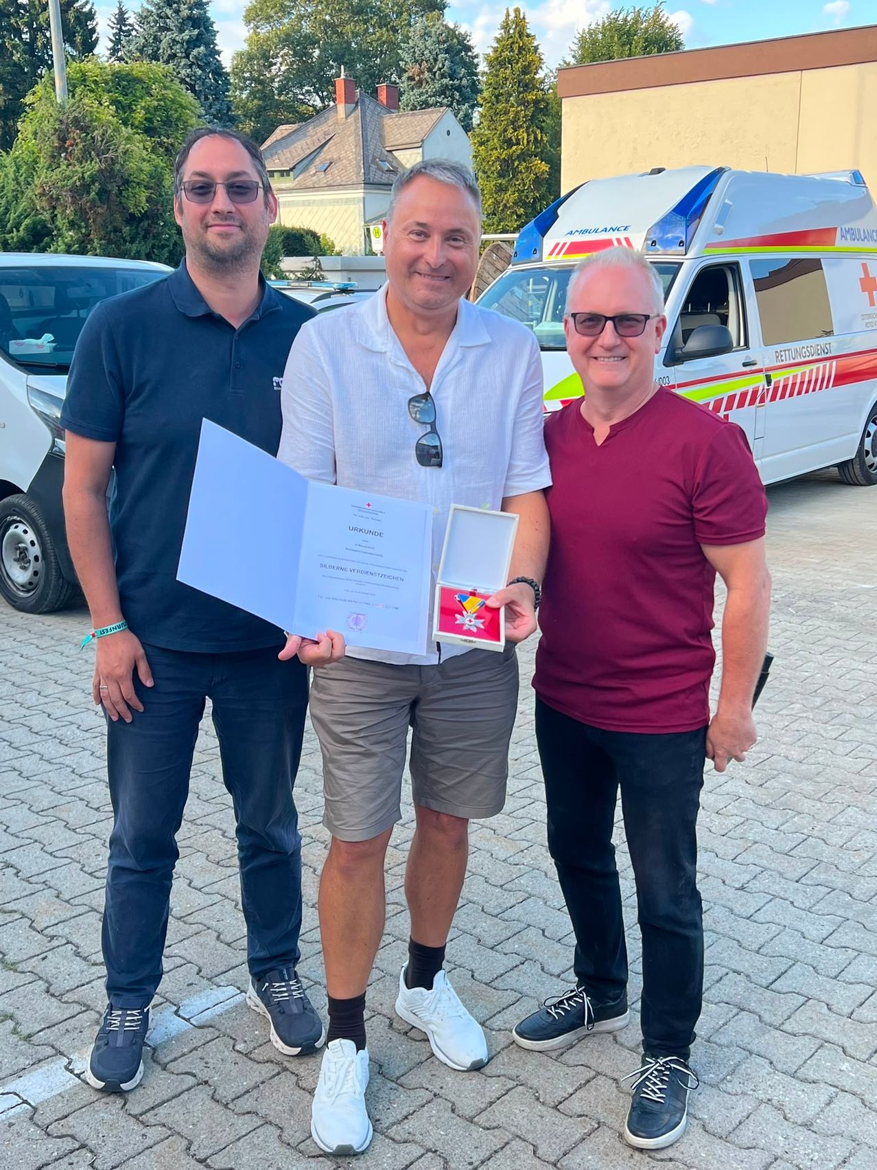 Drei Männer stehen vor einem Krankenwagen, einer hält ein Zertifikat und eine Medaille. Sie lächeln und scheinen eine Errungenschaft zu feiern.