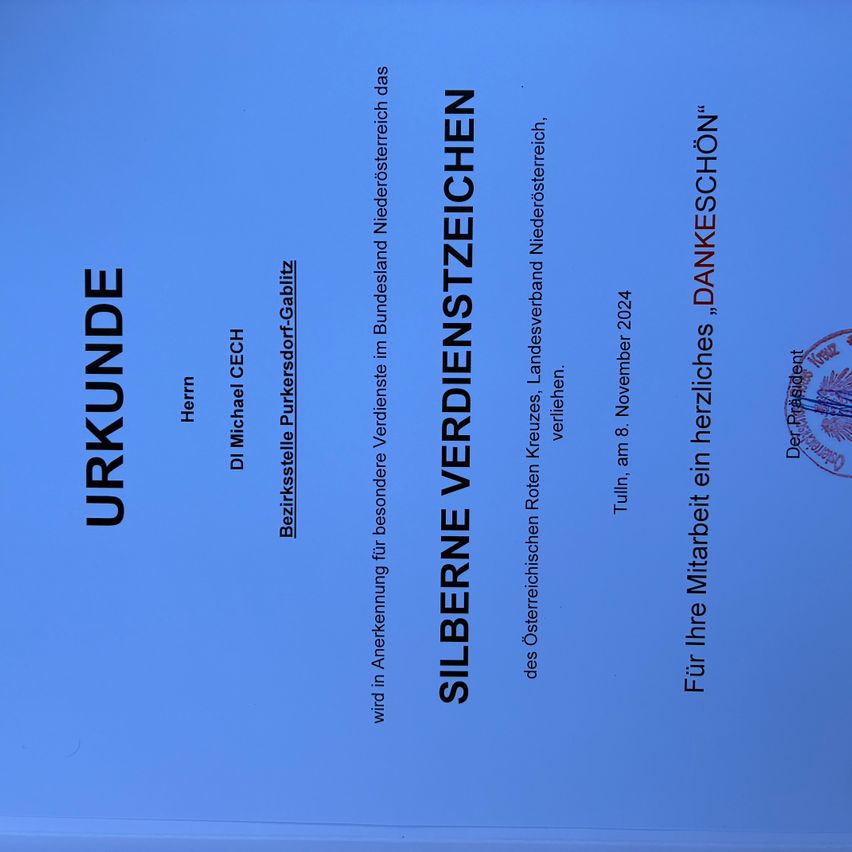 Ein Dokument mit blauem Hintergrund enthält Text auf Deutsch. Es beinhaltet einen Titel 'SILBERNE VERDIENSTZEICHEN', Namen wie 'Michael Pukersdorf-Gabiliz' und ein Datum '8. November 2024'. Auf der rechten Seite befindet sich ein Siegel.