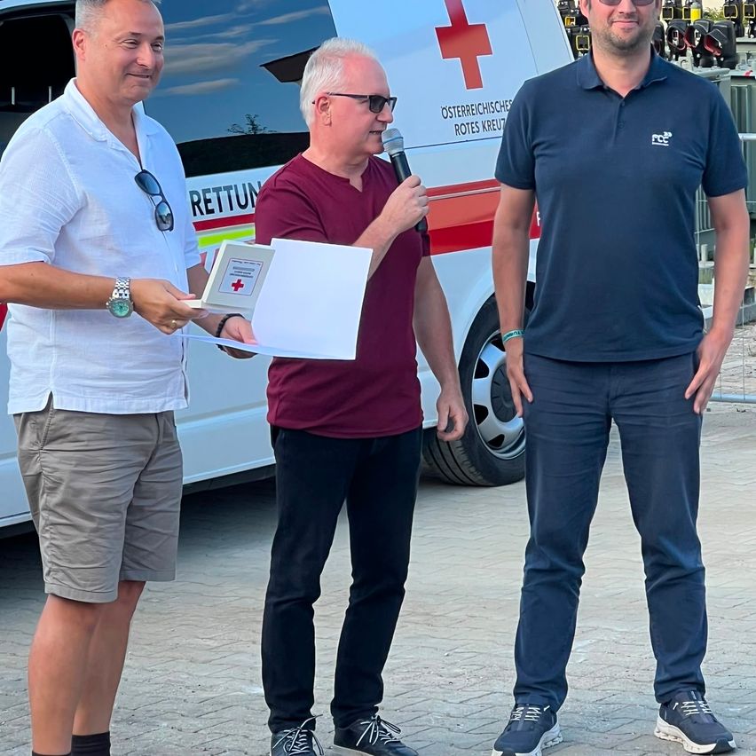 Drei Männer stehen vor einem roten Kreuz-Ambulanz. Der Mann in der Mitte hält ein Mikrofon und ein Papier. Der Mann auf der linken Seite hält ein Papier und einen Stift. Der Mann auf der rechten Seite hat die Hände in den Taschen.
