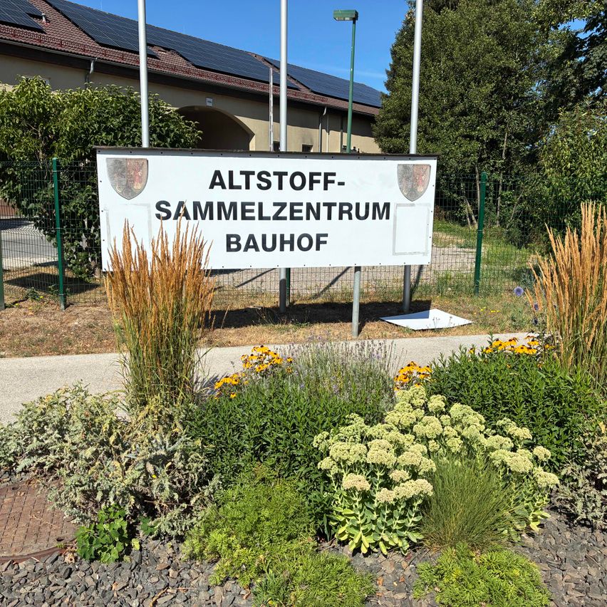 Ein Schild für Altstoff-Sammelzentrum Bauhof steht vor einem Gebäude mit Solarpanelen auf dem Dach.