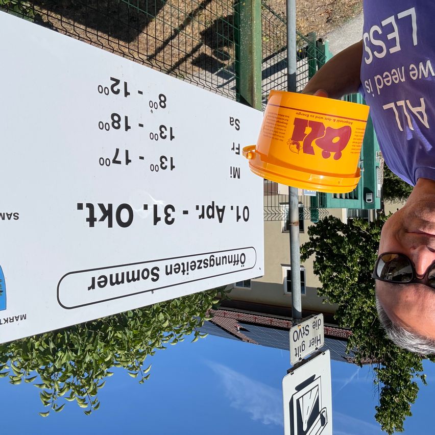 Ein Mann mit einem gelben Eimer mit Logo steht neben einem weißen Schild mit Zahlen und Text. Das Schild hat umgedrehten Text und Zahlen.