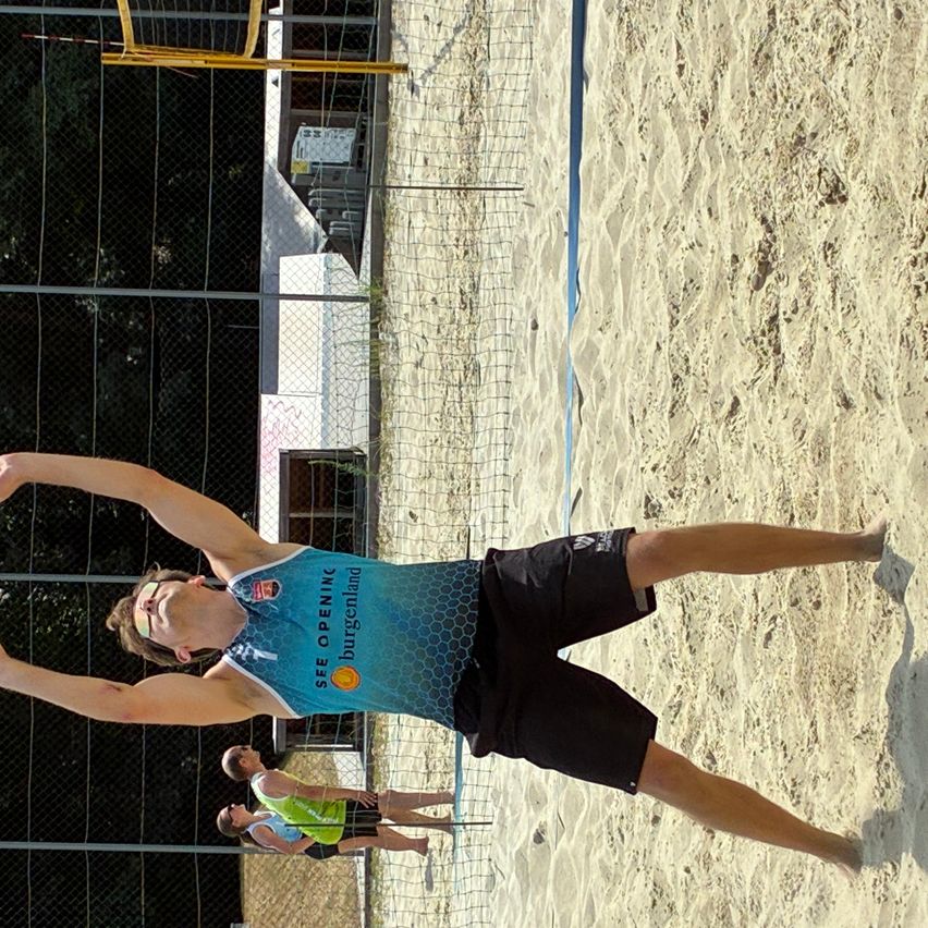 Ein Mann in einem blauen Trikot mit den Worten 'SEE OPENING' springt, um einen Volleyball auf einem sandigen Platz zu schlagen. Dahinter stehen zwei andere Spieler in der Nähe des Netzes.