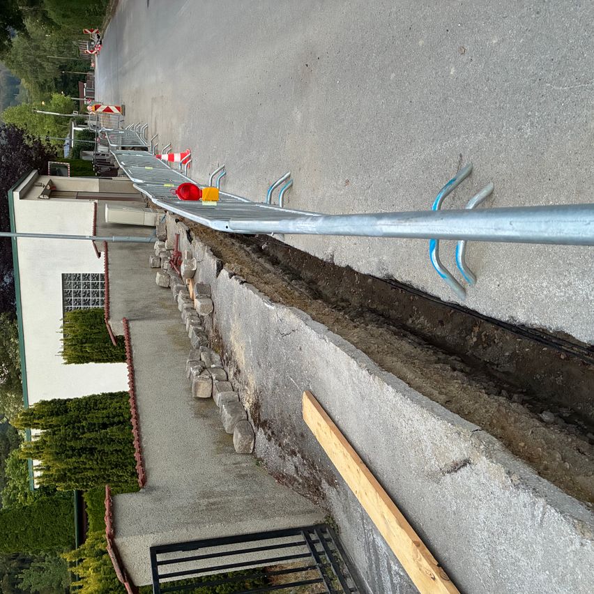 Eine Baustelle mit einer Metallbarriere an einer Straße, neben einem Gebäude mit einer Steinmauer und Grünpflanzen.