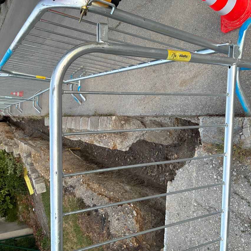 Eine Metallbarriere mit einem gelben Aufkleber ist über einem beschädigten Teil einer Steinmauer platziert, mit einem Verkehrskegel in der Nähe.
