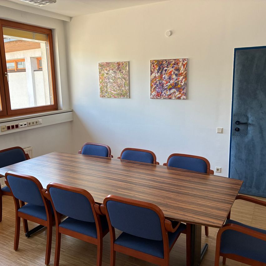 Ein leerer Konferenzraum mit einem Holztisch und blauen Stühlen. Zwei abstrakte Gemälde hängen an der Wand. Ein Fenster mit Jalousien befindet sich links.