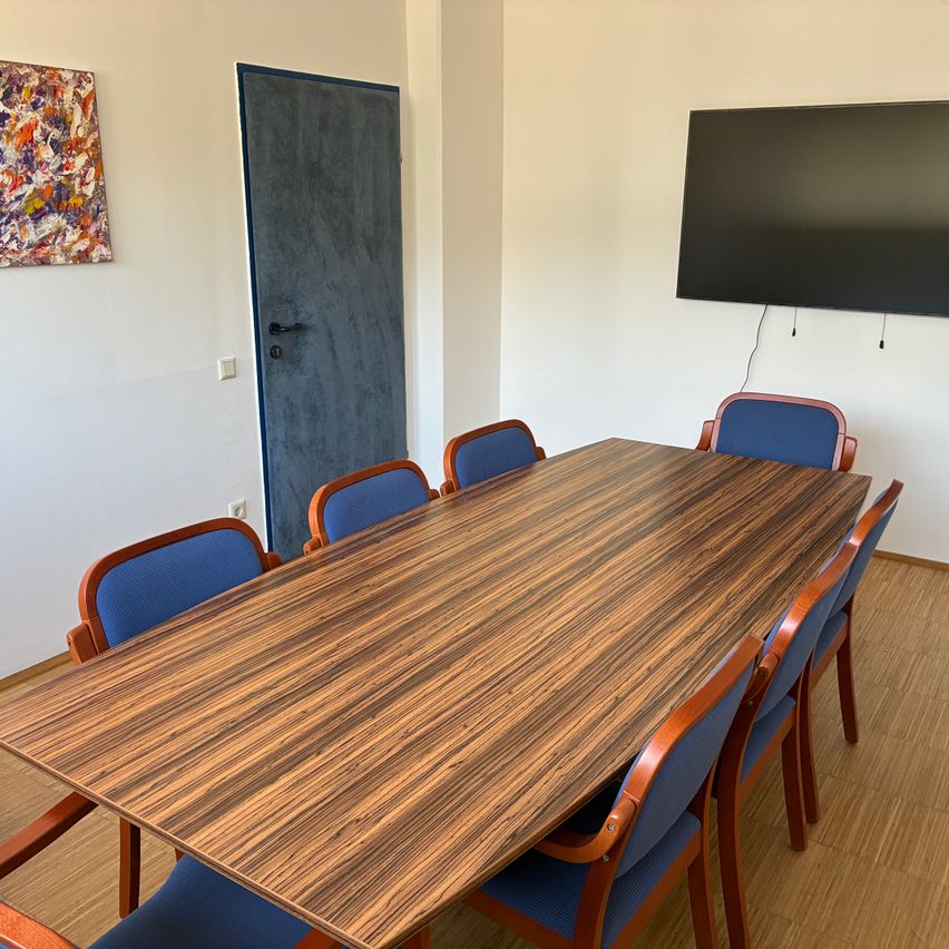 Ein leerer Konferenzraum mit einem Holztisch, blauen Stühlen, einem montierten Fernseher und einer Tür auf der linken Seite.