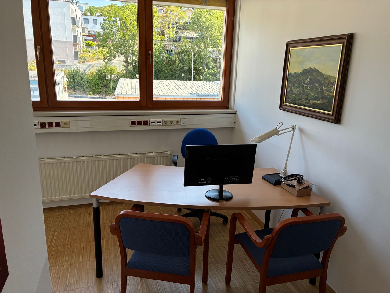 Ein Büro mit einem Schreibtisch, zwei blauen Stühlen, einem Monitor und einer Lampe. An der Wand hängt ein Bilderrahmen und ein Fenster mit Blick auf ein Gebäude und Bäume.