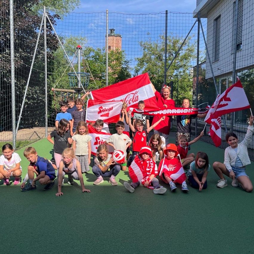 Eine Gruppe von Kindern und Erwachsenen posiert für ein Foto auf einem Sportplatz, halten Flaggen und Banner. Sie tragen rote und weiße Trikots und Mützen. Hinter ihnen steht ein Zaun mit Netz und ein Gebäude.