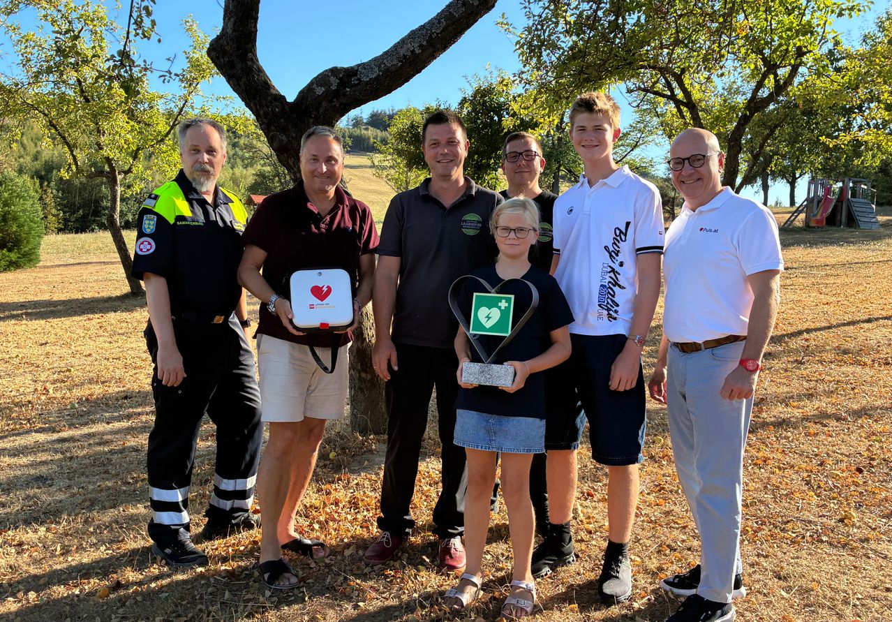 A group of people, including a young girl holding a trophy, stand in a field with trees in the background. The girl has a green heart symbol on her shirt.