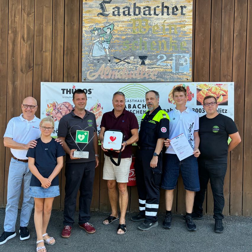 A group of people stand in front of a wooden wall, holding awards and certificates. One man holds a defibrillator. They are smiling and appear to be celebrating an achievement.