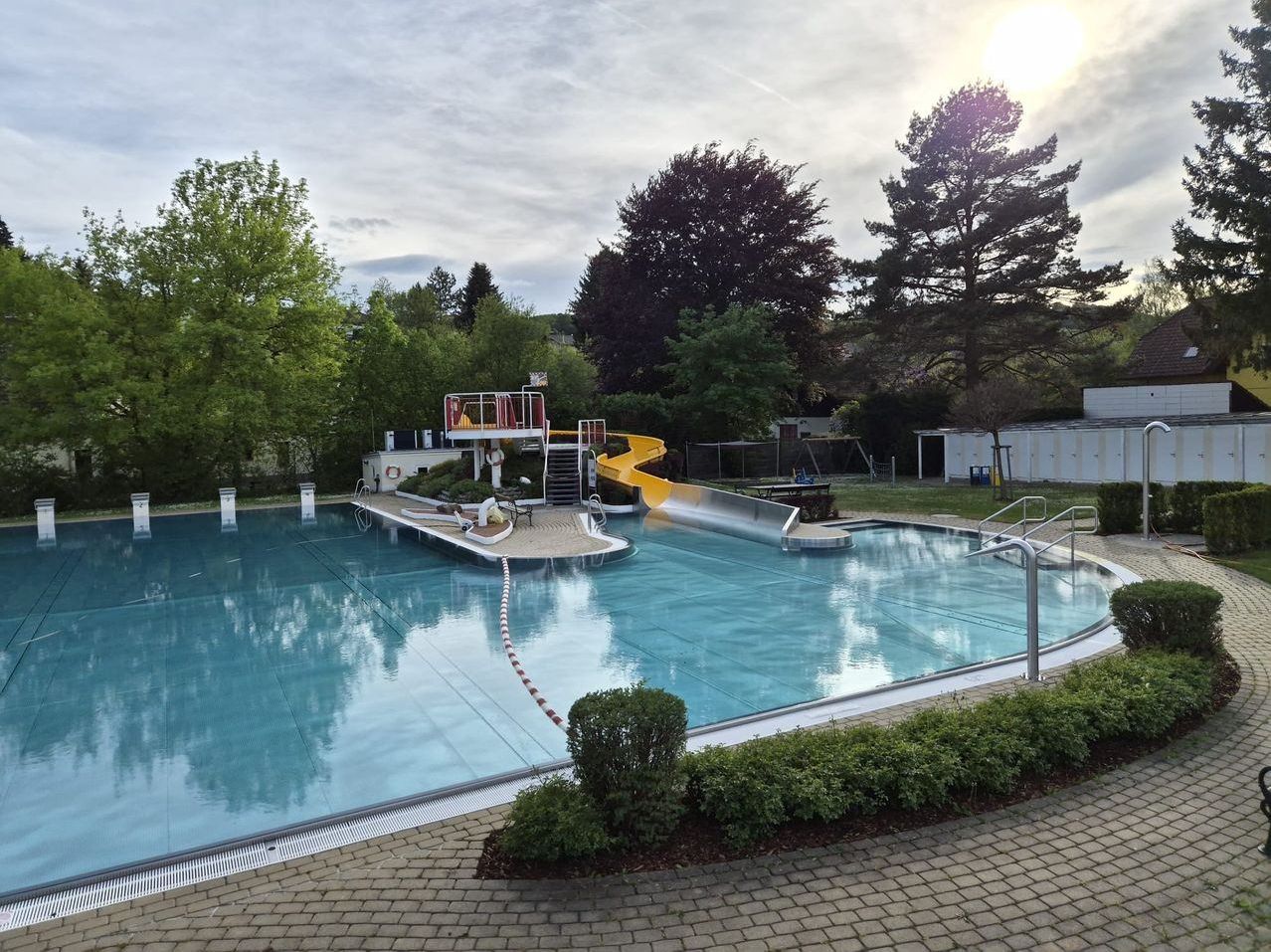 An outdoor swimming pool with a yellow slide and a lifeguard tower, surrounded by greenery and trees, under a cloudy sky.