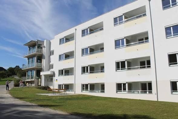 Ein modernes weißes Mehrfamilienhaus mit mehreren Stockwerken, Balkonen und einem Rasen davor. Eine Bank und Menschen sind in der Nähe des Eingangs.