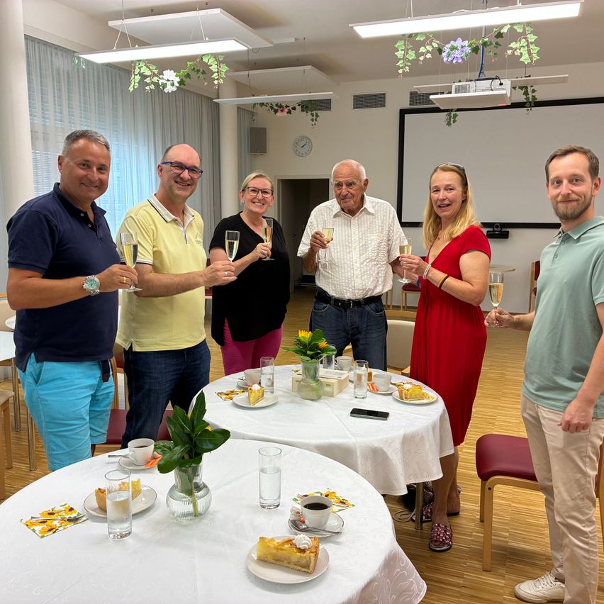 Eine Gruppe von Menschen steht um einen Tisch mit Essen und Getränken, lächelt und hält Weingläser. Es gibt zwei Tische mit weißen Tischdecken, einer mit einer Blumenarrangement und der andere mit einem Teller Essen. Der Raum hat eine Projektionsleinwand und eine Uhr an der Wand.