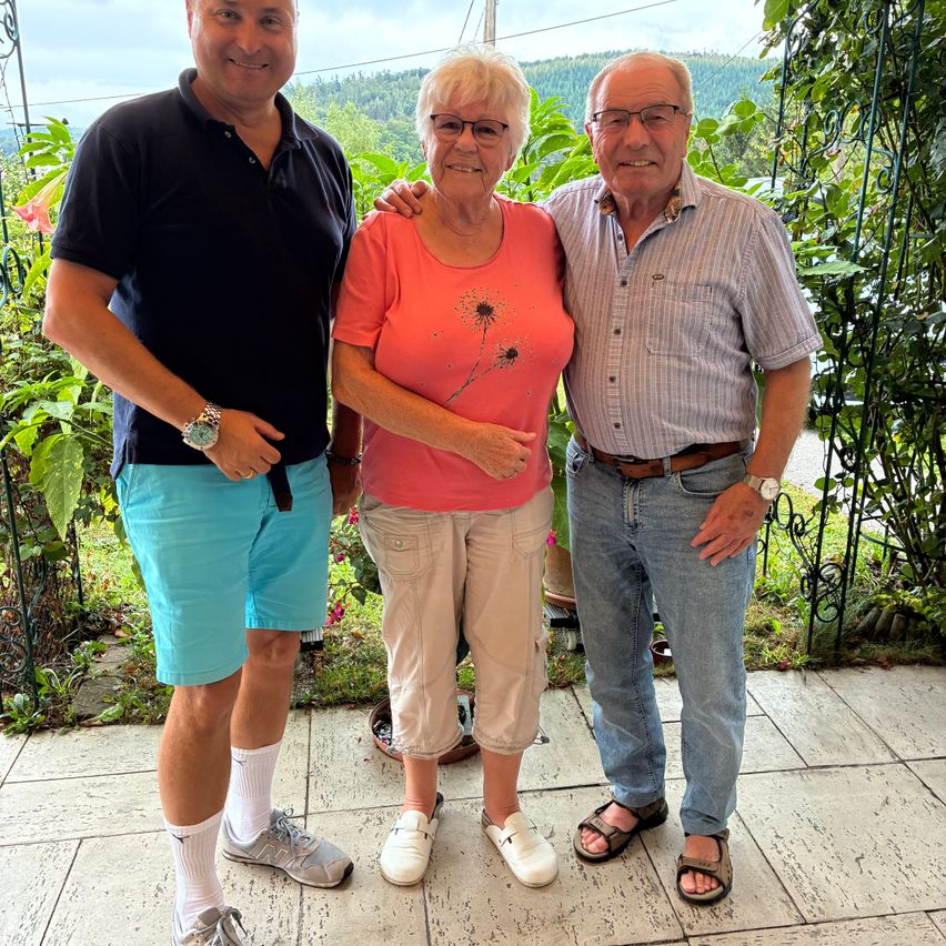 Drei Menschen stehen zusammen in einem Garten und lächeln für ein Foto. Der Mann links trägt ein schwarzes Shirt und blaue Shorts. Die Frau in der Mitte trägt ein rosa Shirt und Khaki-Hosen. Der Mann rechts trägt ein gestreiftes Hemd und Jeans.