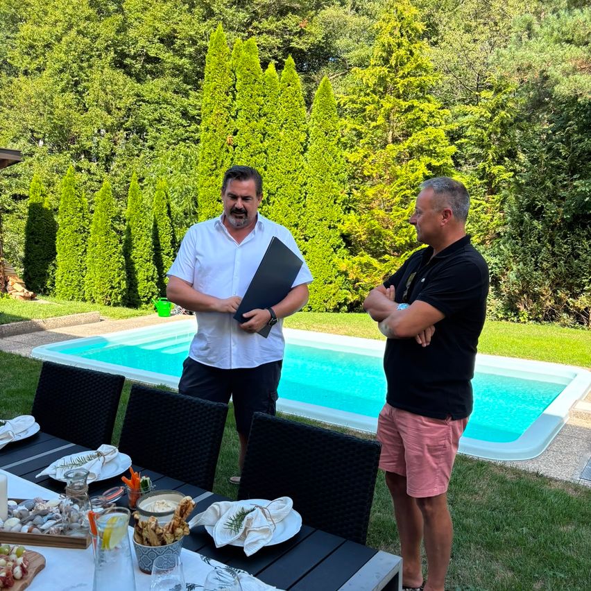Zwei Männer stehen im Garten neben einem Pool, einer hält eine Mappe.