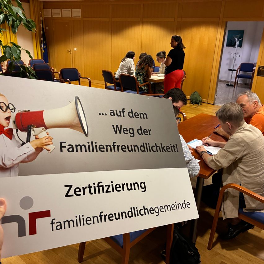 Ein von jemandem gehaltenes Plakat zeigt ein Kind, das einen Megafon mit dem Text '... auf dem Weg der Familienfreundlichkeit! Zertifizierung familienfreundlichegemeinde' hält. Im Hintergrund sitzen Menschen an einem Tisch, möglicherweise in einer Besprechung.