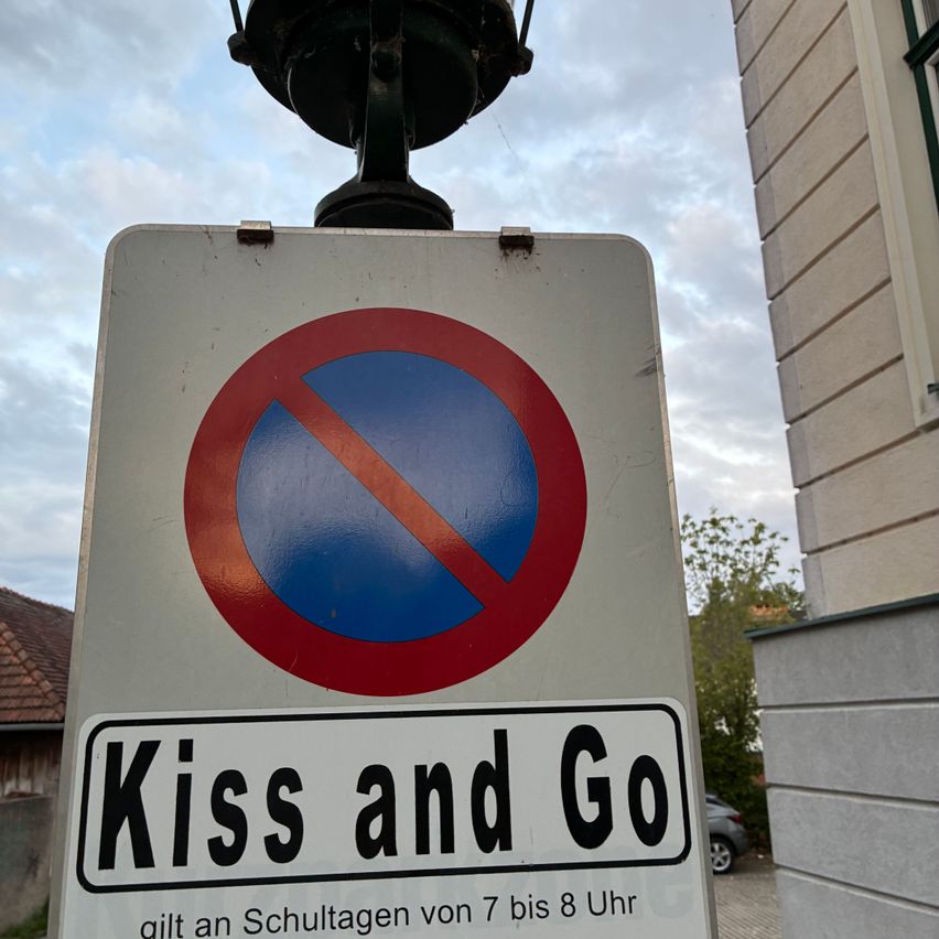 Ein weißes Schild mit einem roten Kreis und einem Strich, darunter steht 'Kiss and Go' und ein Zeitbereich von 7 bis 8 Uhr.