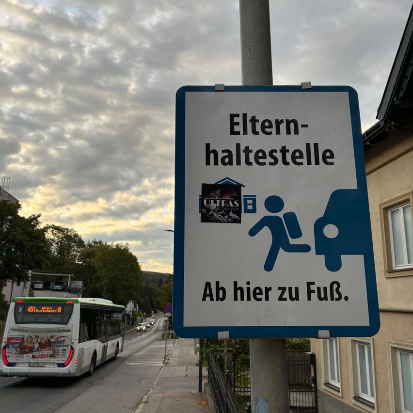 Ein Schild an einem Pfosten weist auf eine Eltern-Kind-Haltestelle hin, mit einer laufenden Person und einem Auto. Das Schild ist an einem Pfosten in der Nähe eines Busses und eines Gebäudes angebracht.