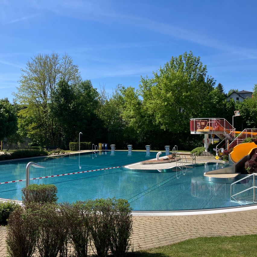 Ein Freibad mit einer roten Rutsche und einer Sprungplattform an einem sonnigen Tag. Bäume und Büsche umgeben den Pool.