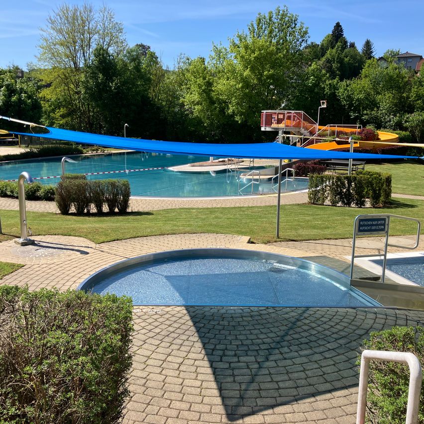 Ein Freibad mit einem blauen Sonnensegel und einer Wasserrutsche im Hintergrund. Der Pool ist von Grünflächen und gepflasterten Wegen umgeben.