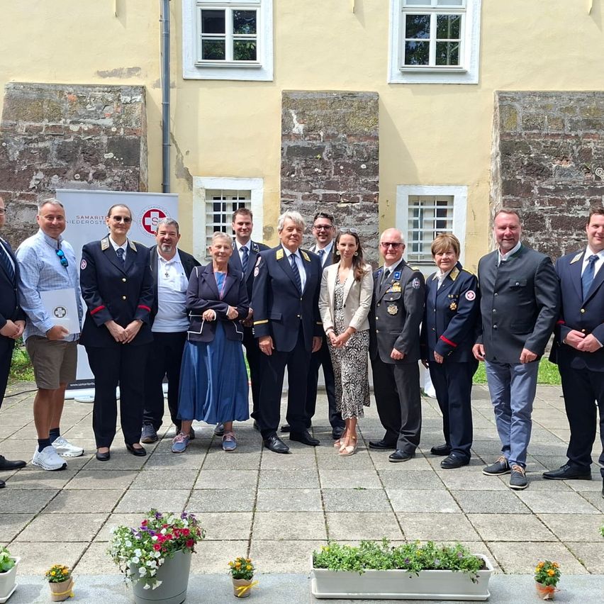 A group of people dressed in formal attire stand in front of a building with brick walls and white windows. There are potted plants in front.
