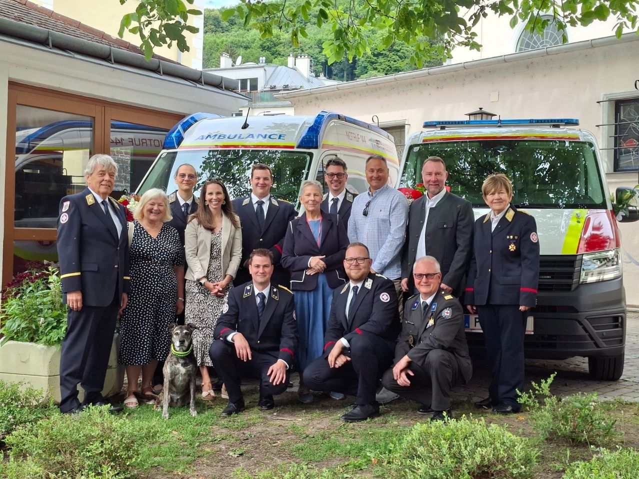 A group of people in uniforms and suits stand in front of ambulances, smiling and posing for a photo. A dog is sitting in front of them.