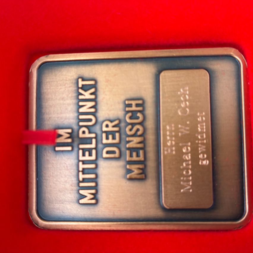 A silver plaque with golden text that reads 'IM MITTELPUNKT DER MENSCH'. It features a name 'Michael W.' and an additional text 'Frieden' on the right.