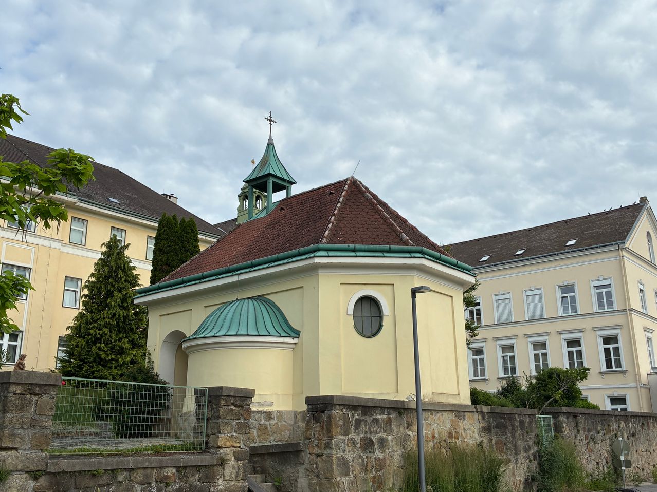 A small church with a brown roof and green accents. It features a bell tower with a cross, a window with a round frame, and is surrounded by a stone wall.