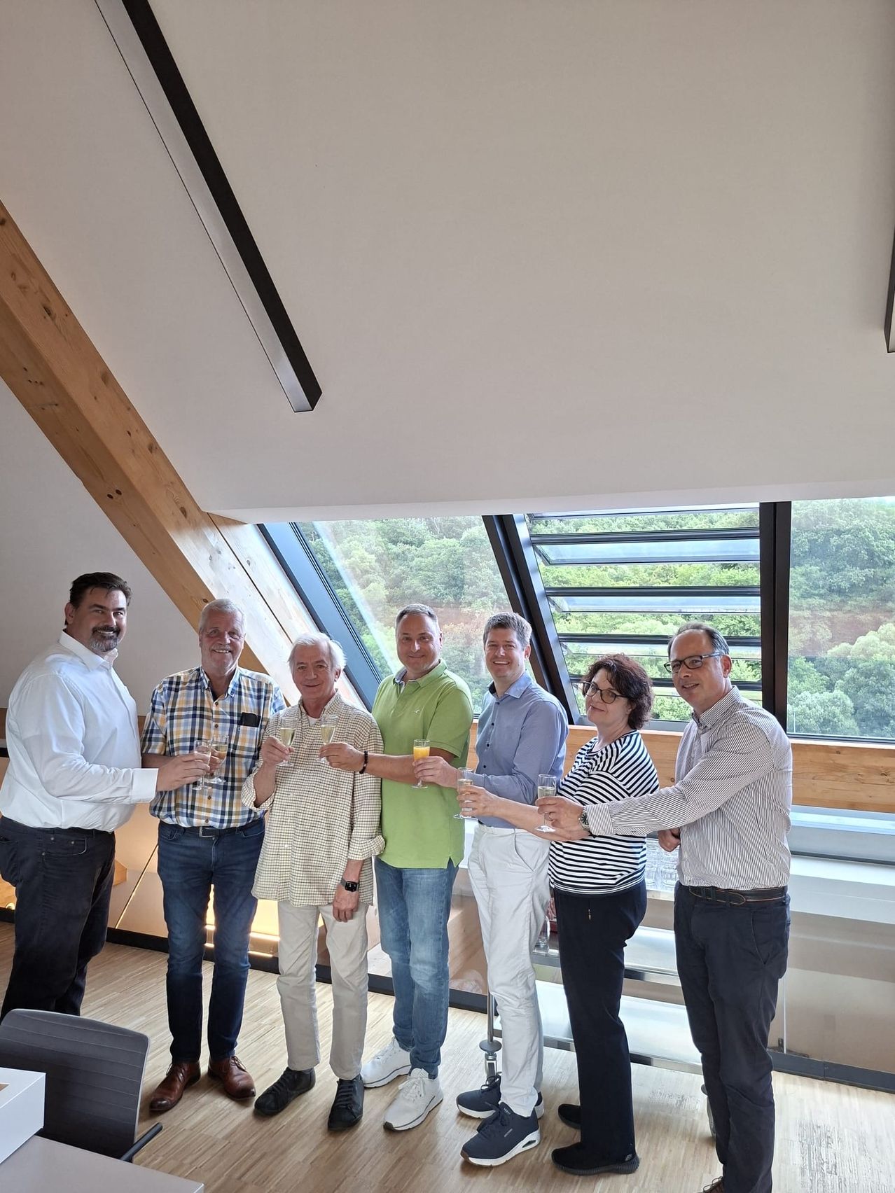 A group of six adults stands inside a room, holding wine glasses, likely celebrating. They are positioned near a window that overlooks a forested area.
