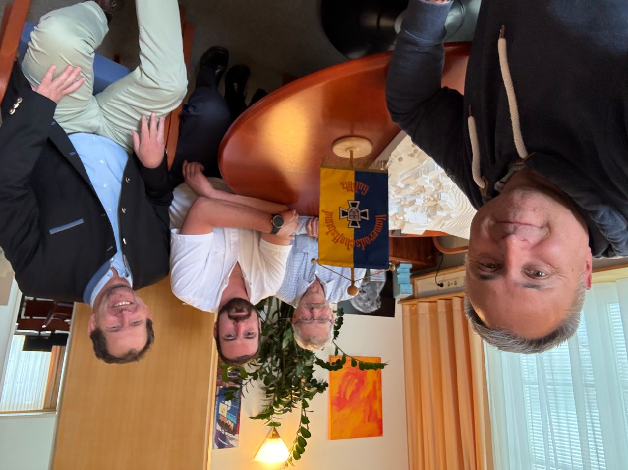Fünf Männer versammeln sich in einem Raum. Ein Mann sitzt auf dem Tisch, und der andere liegt auf dem Boden. Eine Flagge mit Text und Kreuz ist am Tisch befestigt.