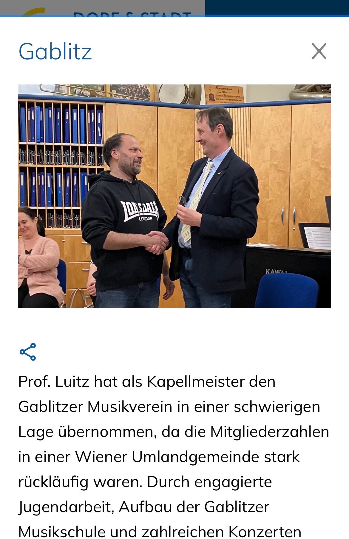 Zwei Männer schütteln Hände in einem Raum mit Holzschränken, einem Klavier und einer sitzenden Frau. Ein Mann trägt Krawatte und Anzug, der andere eine Kapuzenjacke.