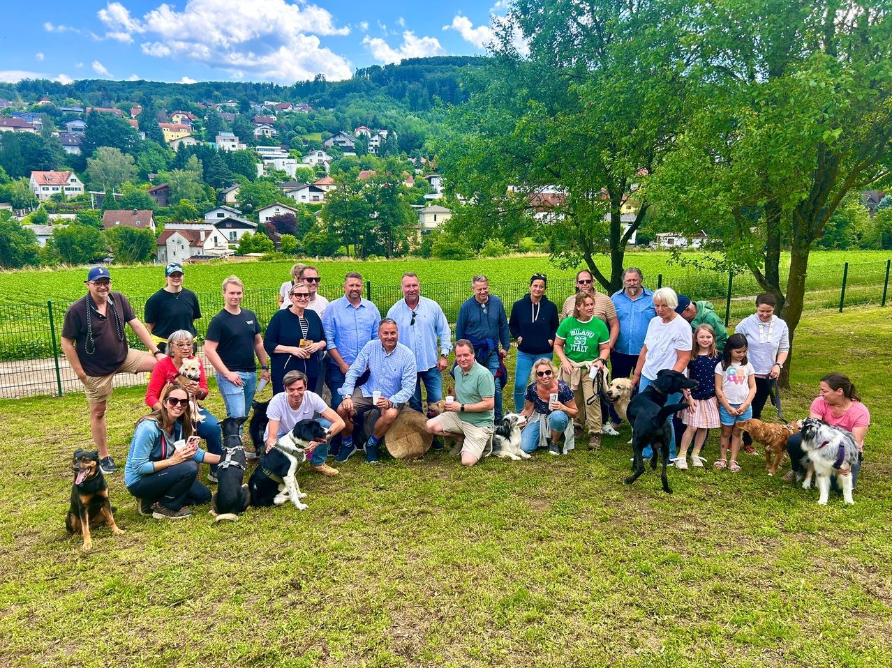 Eine Gruppe von Menschen und ihren Hunden posiert für ein Foto auf einer Wiese mit einem Dorf und Bergen im Hintergrund.