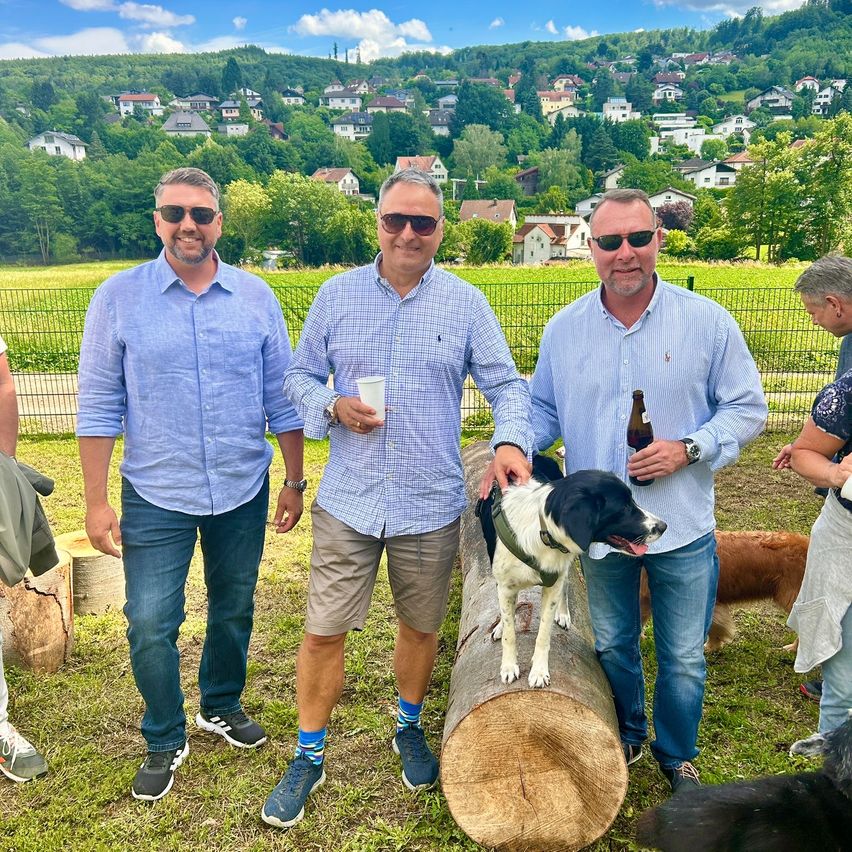 Drei Männer posieren auf einem Feld, stehen auf einem Baumstamm, einer hält einen Hund. Ein Mann in der Mitte hält einen Becher. Hinter ihnen ist ein Zaun, und mehr Menschen und Hunde sind in der Nähe.