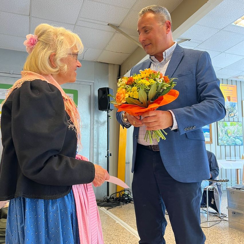 Ein Mann mit einem Blumenstrauß und eine Frau mit einem rosa Schal stehen in einem Raum.