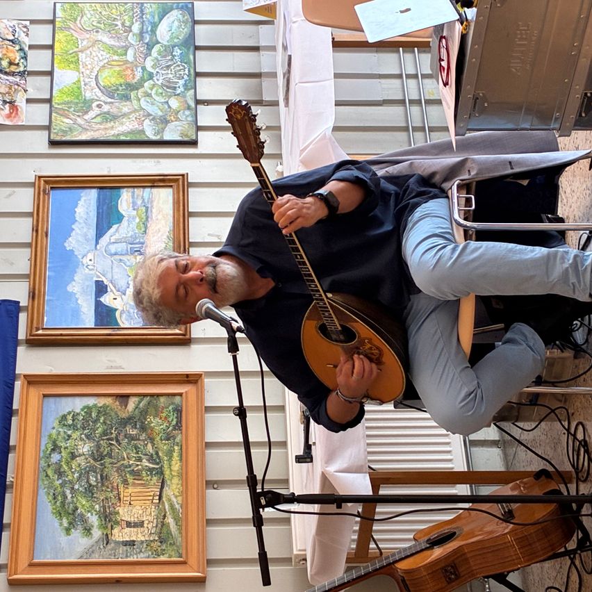 Ein älterer Mann spielt eine Mandoline vor einem Mikrofon und drei Gemälden.