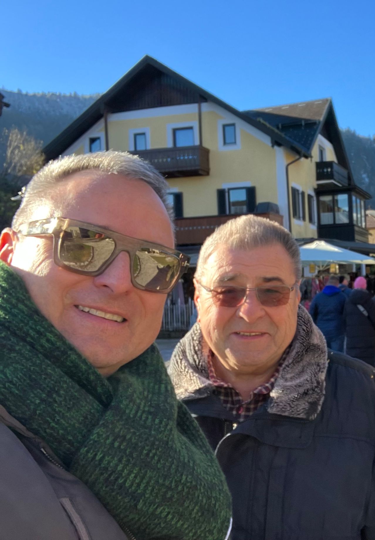Zwei Männer lächeln vor einem gelben Haus mit Bergen im Hintergrund. Beide tragen Sonnenbrillen und Schals.