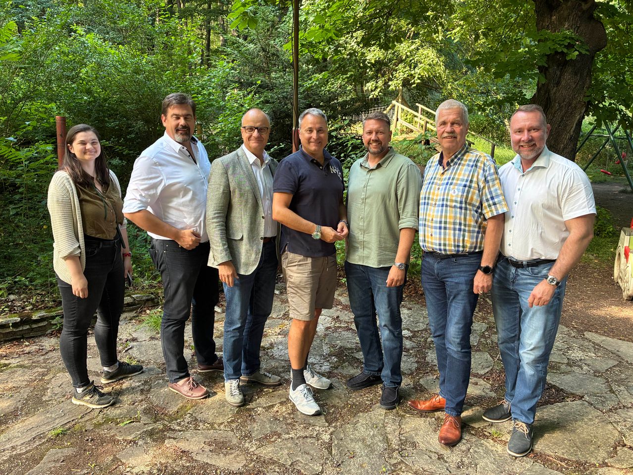 Eine Gruppe von Männern und einer Frau steht in einem Wald und lächelt für ein Foto. Die Frau trägt ein Armband. Hinter ihnen befindet sich eine Holzbrücke.