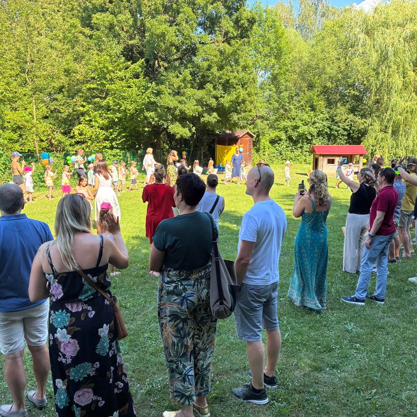 Eine Gruppe von Menschen steht an einem sonnigen Tag in einem Park. Einige machen Fotos von spielenden Kindern. Dahinter ist ein kleines Spielhaus zu sehen, und Bäume bilden eine natürliche Kulisse.