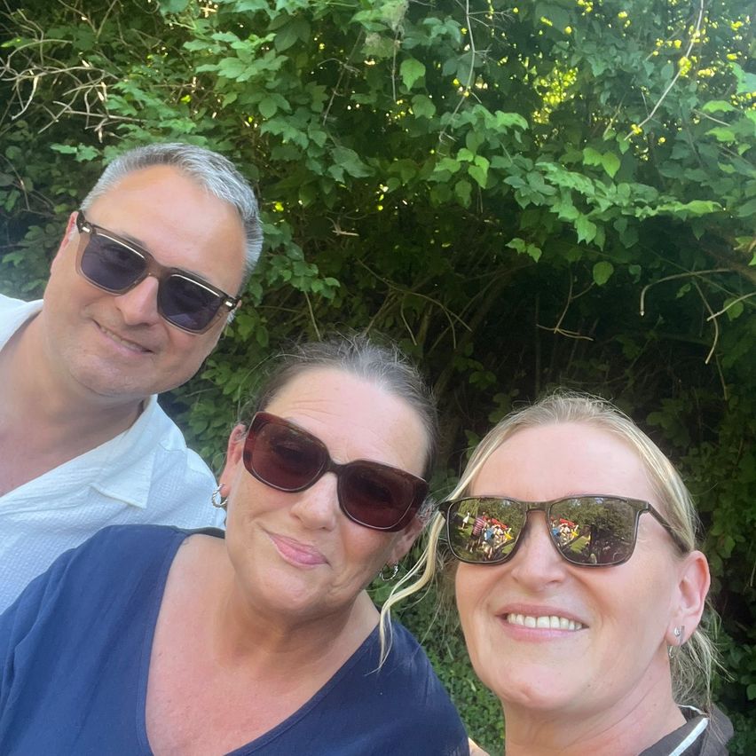 Drei Personen lächeln und posieren für ein Foto mit grüner Vegetation im Hintergrund. Einer ist ein Mann in einem weißen Hemd, eine weitere eine Frau in einem blauen Hemd und die dritte eine Frau mit blonden Haaren.