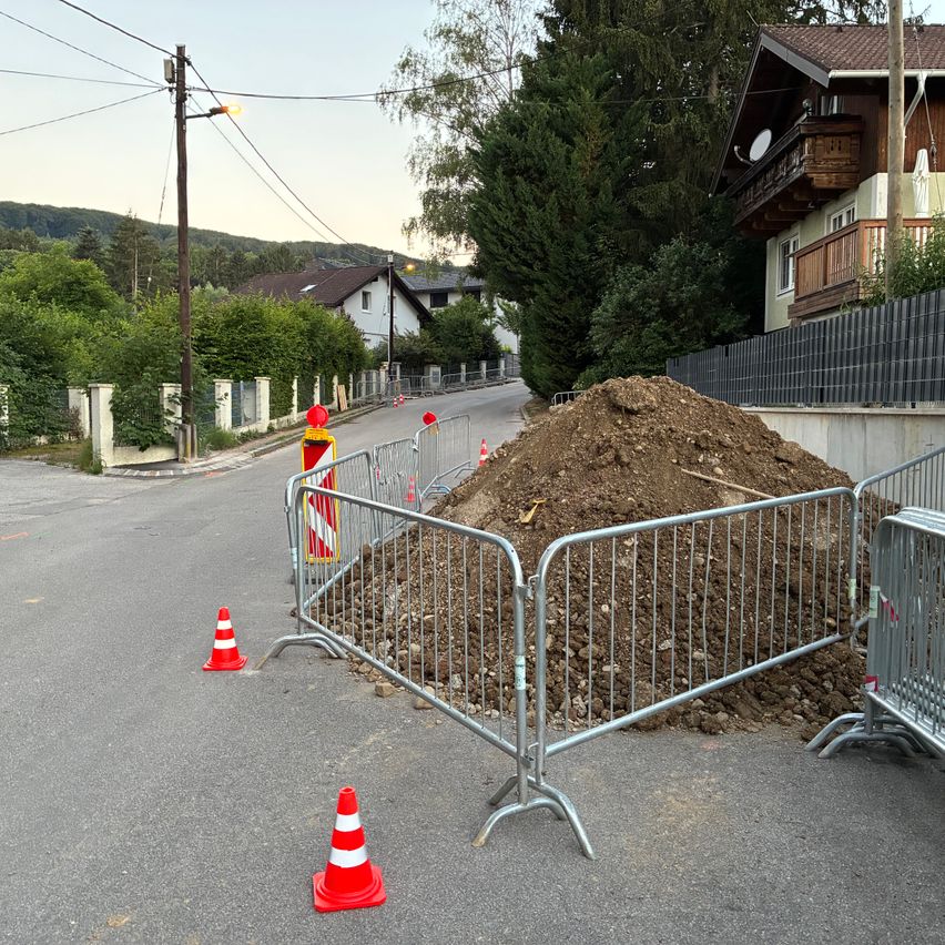 Eine Straße mit Metallbarrieren und orangefarbenen Verkehrskegel rund um einen großen Haufen Erde. Ein Haus ist auf der rechten Seite.