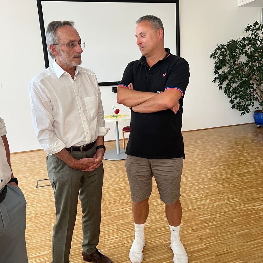 Zwei Männer in einem Raum, einer in einem schwarzen Shirt mit verschränkten Armen und der andere in einem weißen Hemd mit verschränkten Händen. Dahinter steht ein kleiner Tisch mit einer Blume und einer Tafel.