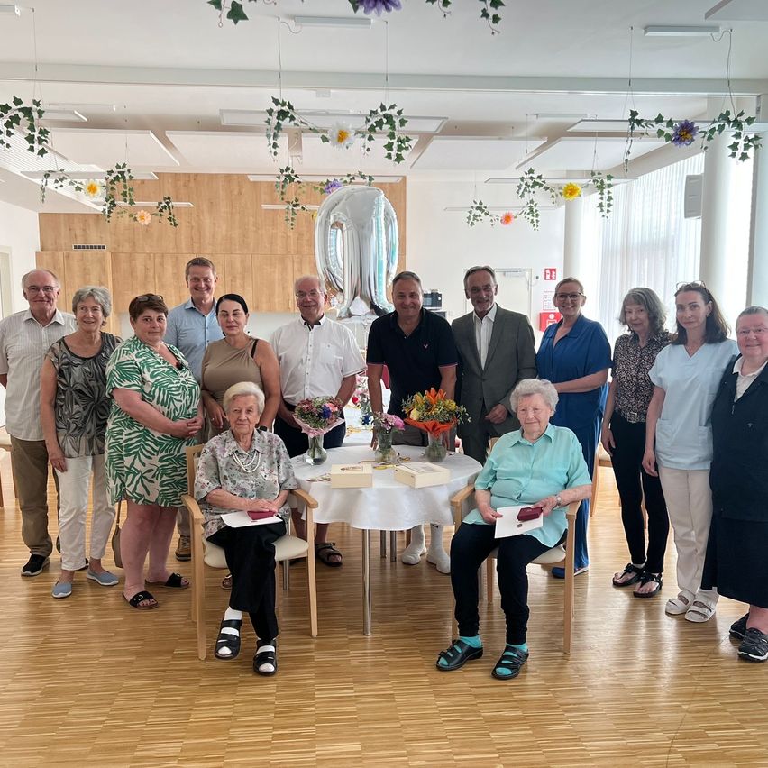 Eine Gruppe von Menschen versammelt sich in einem Raum zur Feier, wobei zwei ältere Frauen sitzend Blumen erhalten. Ein Tisch mit Kuchen und Geschenken ist von stehenden Personen umgeben. Blumendekorationen zieren die Decke.