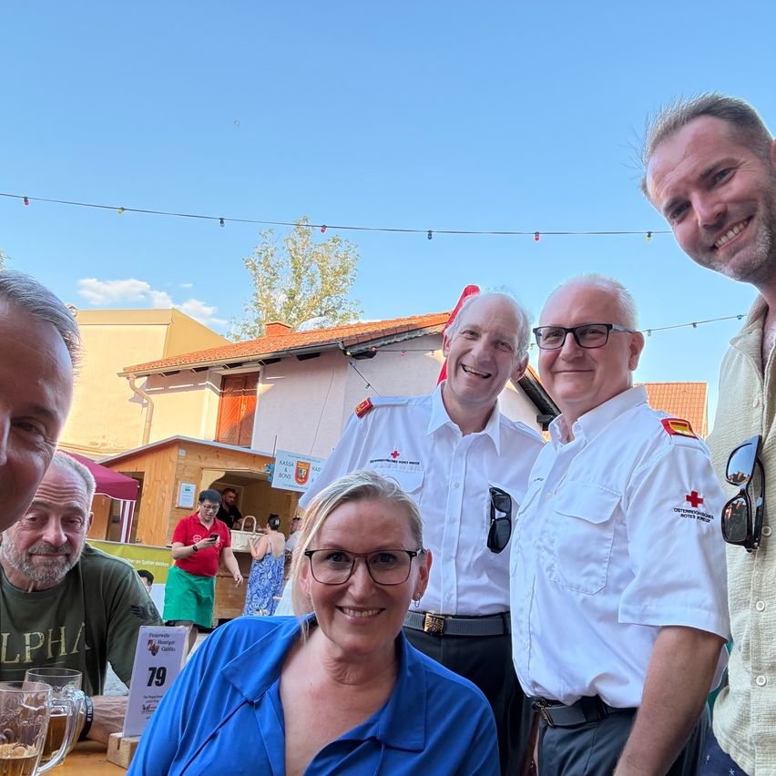 Mehrere Menschen lächeln für ein Foto. Zwei Männer tragen weiße Uniformen mit roten Kreuzen. Eine Frau in einem blauen Shirt lächelt vor ihnen. Ein Mann in einem grünen Shirt sitzt an einem Tisch mit zwei Biergläsern. Im Hintergrund steht ein Haus mit einem roten Dach und Bäumen.
