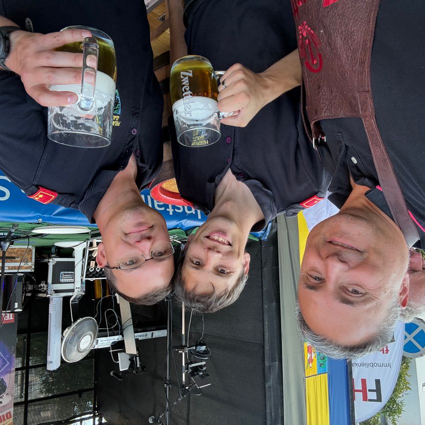 Drei Männer mit Bierkrügen lächeln in die Kamera. Einer trägt eine Brille, ein anderer ein braunes Weste. Der Hintergrund zeigt ein blaues und gelbes Banner.