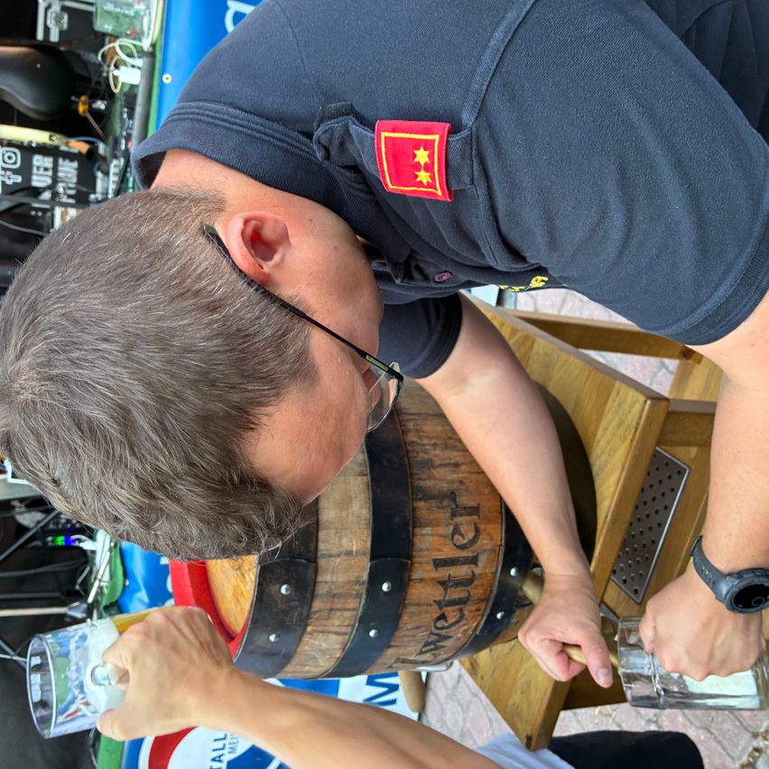 Ein Mann mit Brille und blauem Shirt mit rotem Aufnäher schenkt ein Getränk aus einem Holzfass in ein Glas ein.