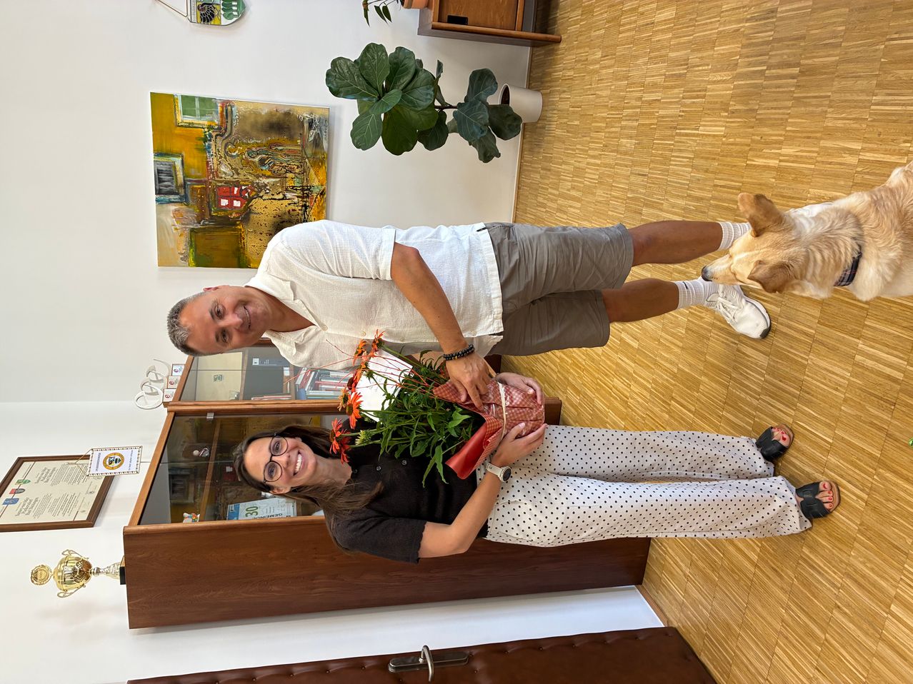 Ein Mann und eine Frau stehen lächelnd im Innenraum. Der Mann hält einen Blumenstrauß. Hinter ihnen ruht ein Hund auf dem Boden. Ein Topf mit Pflanze, ein Gemälde und ein Regal mit Büchern im Hintergrund.