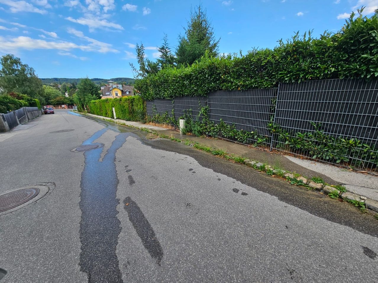 Eine Straße mit einer nassen Stelle auf der Straße, einem Metallzaun und einem gelben Haus in der Ferne.